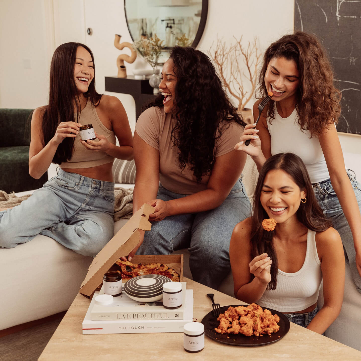 A group of 4 smiling women with dark hair, sit near each other in a living room, enjoying pizza and wings. One holds a bottle of arrae capsules