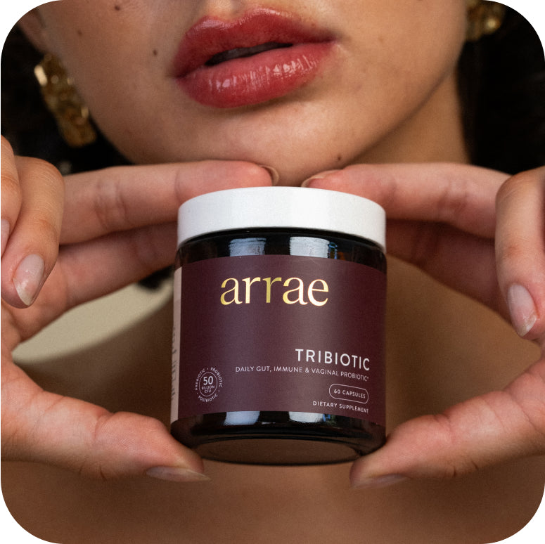 A woman is holding a jar of Arrae Tribiotic supplement, showing the label clearly. The jar is in focus with her hands framing it, and the background is blurred, drawing attention to the product.