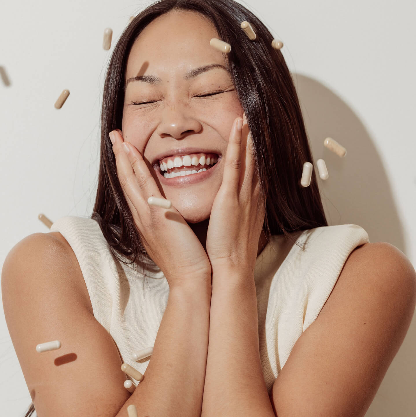 A woman smiles, and holds her cheeks while arrae capsules fall around her like confetti