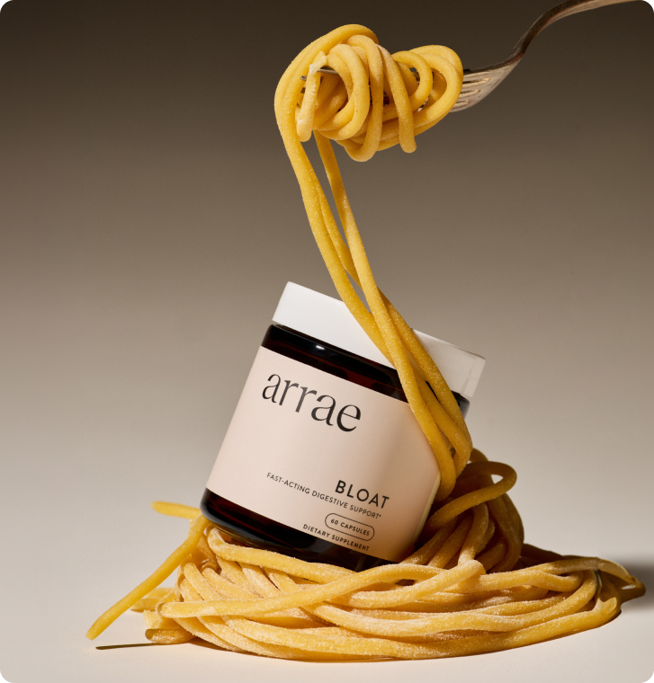 A jar of Arrae Bloat supplement is surrounded by spaghetti noodles, with some noodles artistically spiraling around a fork above the jar. The jar's label reads Bloat - Fast-Acting Digestive Support, and the image has a neutral, minimalistic background.